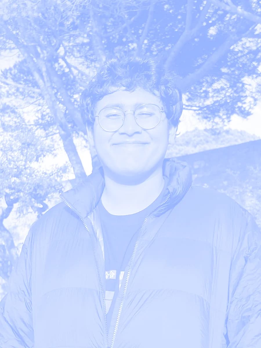 Vidush, smiling, in glasses and a dark puffer jacket, under pine trees